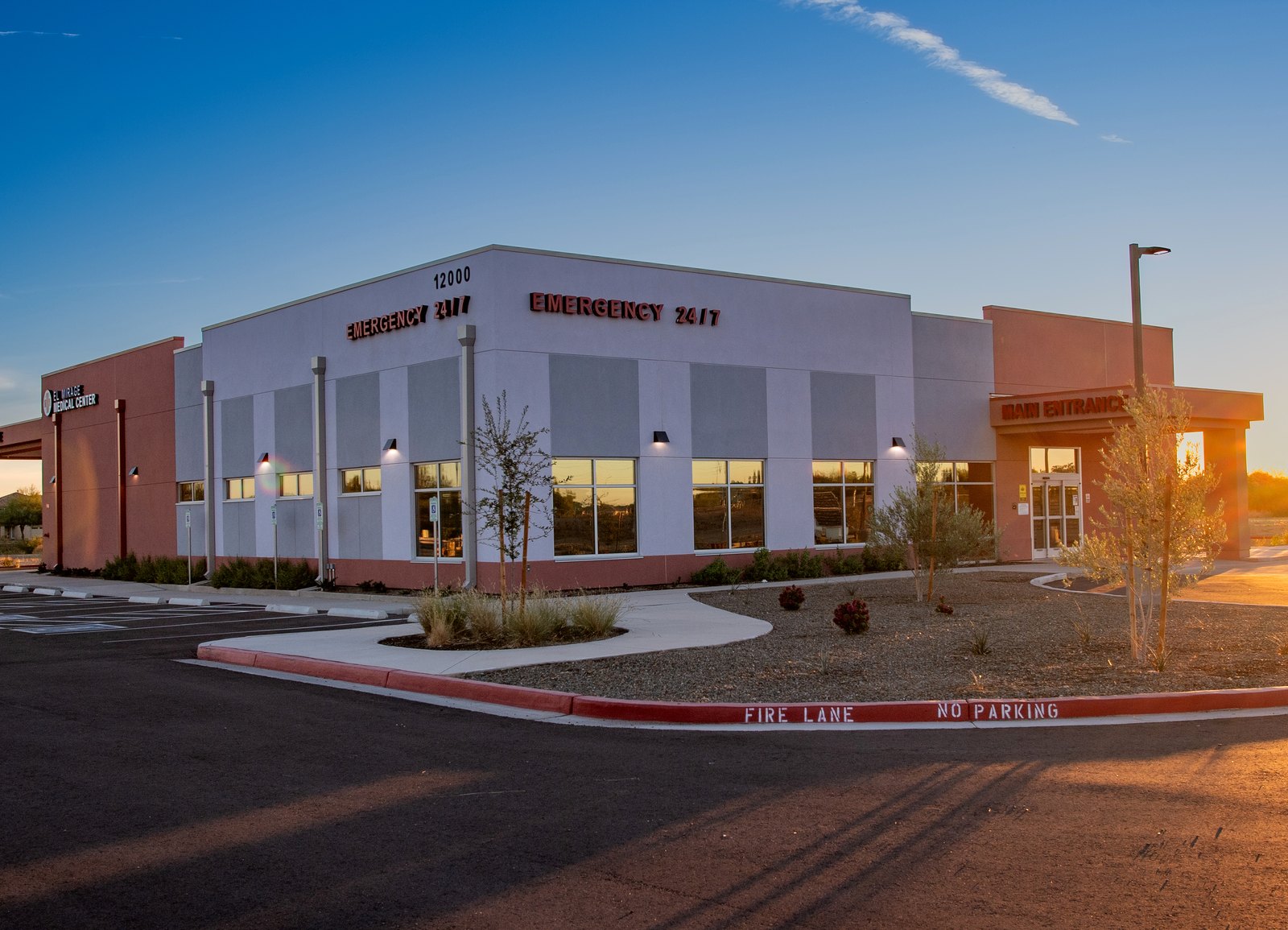 The El Mirage ER building seen from the car park at dusk, with EMERGENCY 24/7 and the street number 12000 lit on the parapet and the Main Entrance canopy at the right.