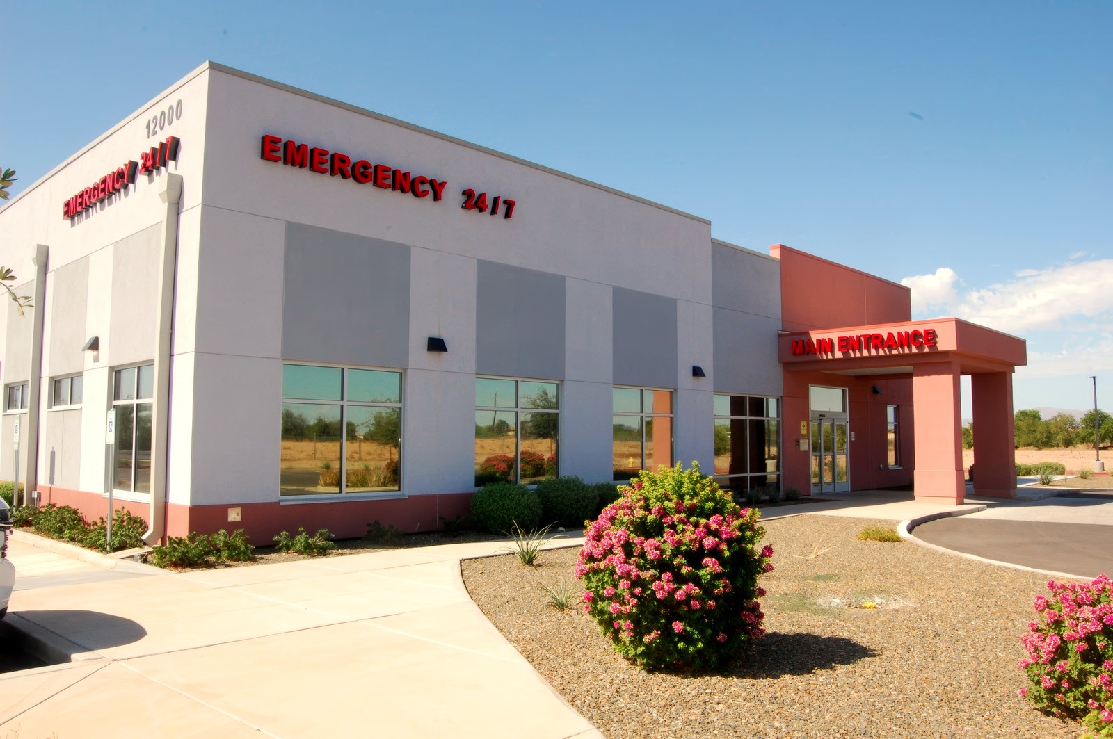 El Mirage ER seen from the car park — EMERGENCY 24/7 on the parapet, the street number 12000, and the red main entrance canopy.