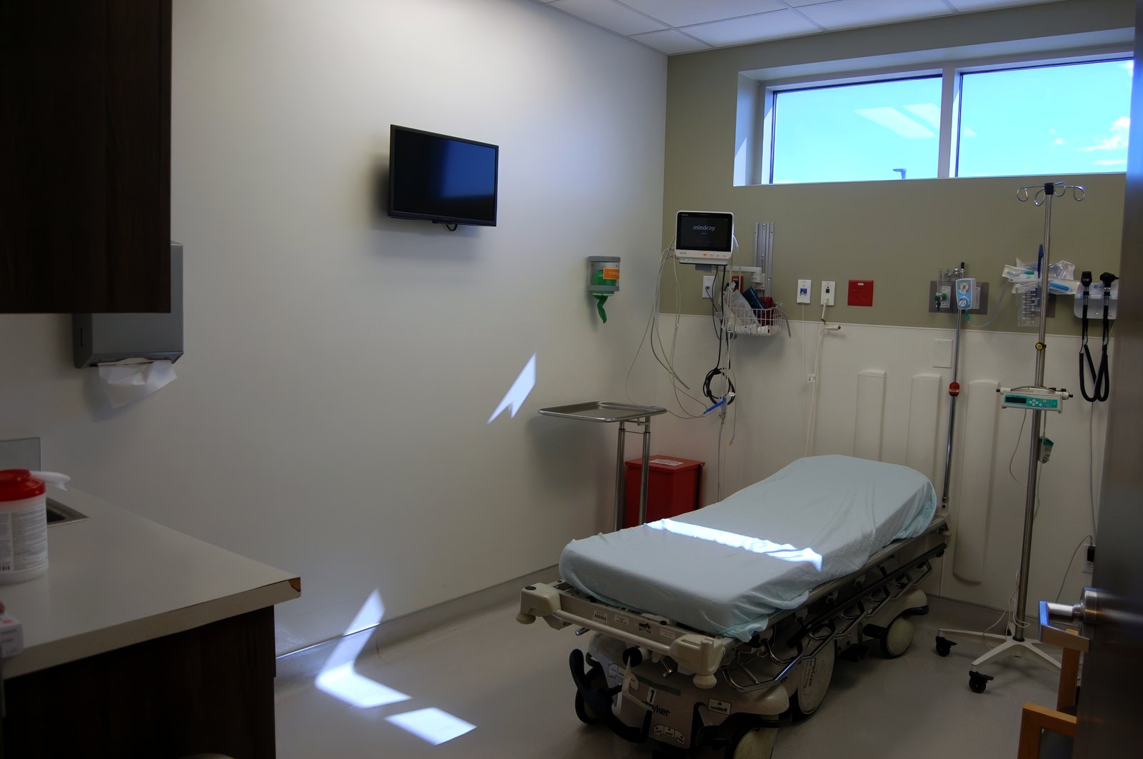 An empty treatment bay at El Mirage ER, with a made-up gurney, a wall-mounted monitor, and a window letting in daylight.