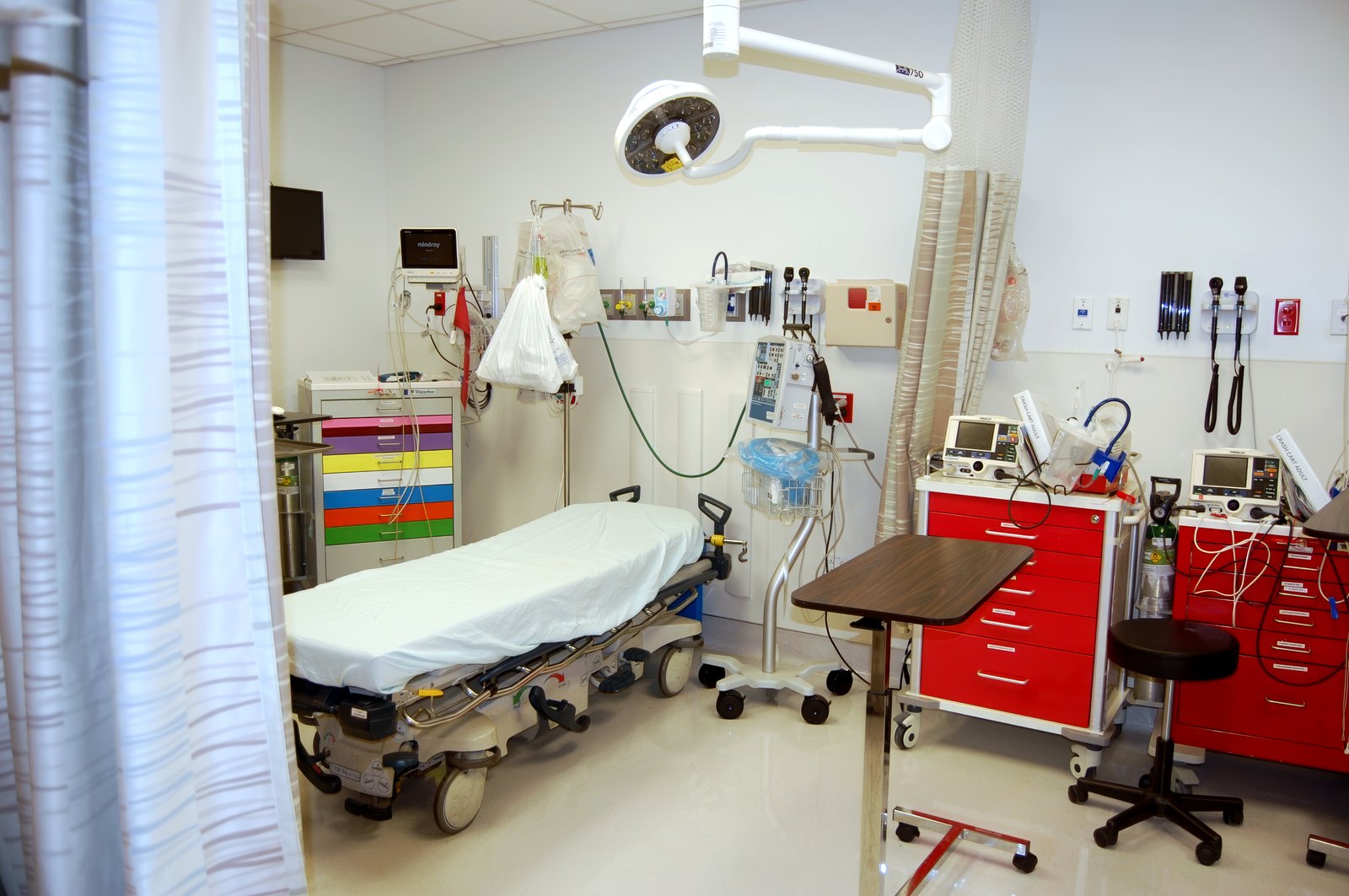 An empty treatment room at El Mirage ER — a gurney beneath an overhead procedure light, a crash cart, monitors, and diagnostic instruments mounted on the wall.
