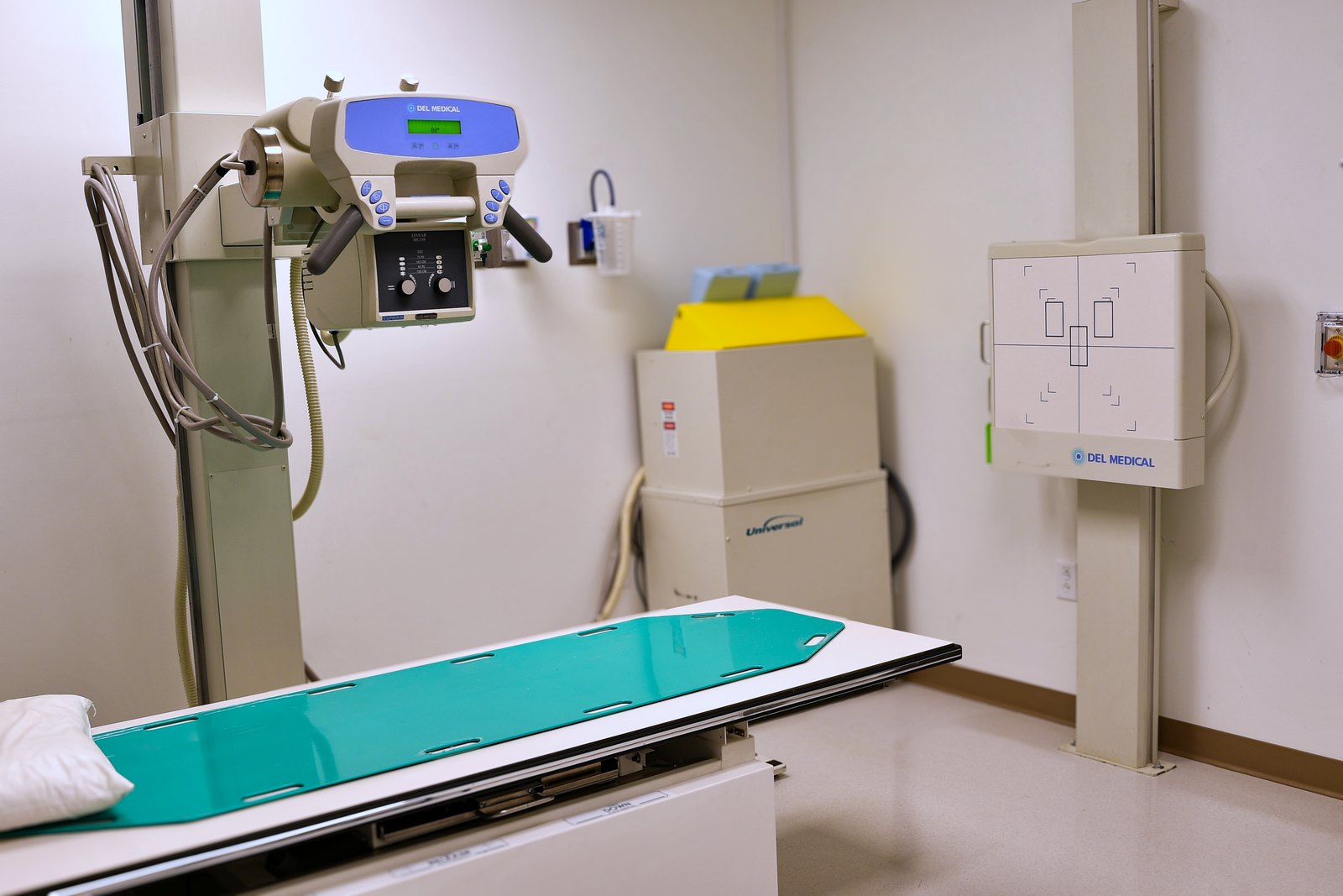 An X-ray room at El Mirage ER, with a column-mounted tube head, a green-topped patient table, and a wall-mounted detector panel.
